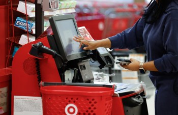 A person using a self-checkout kiosk in a Target store, touching the screen with one hand while holding a phone in the other.