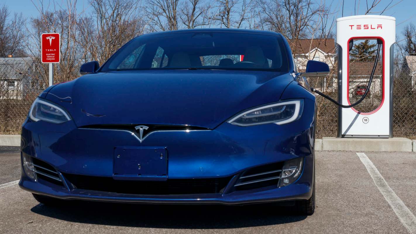 A blue Tesla car is parked at a charging station with a clear blue sky in the background.