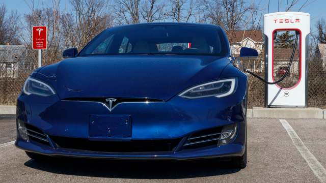 A blue Tesla car is parked at a charging station with a clear blue sky in the background.