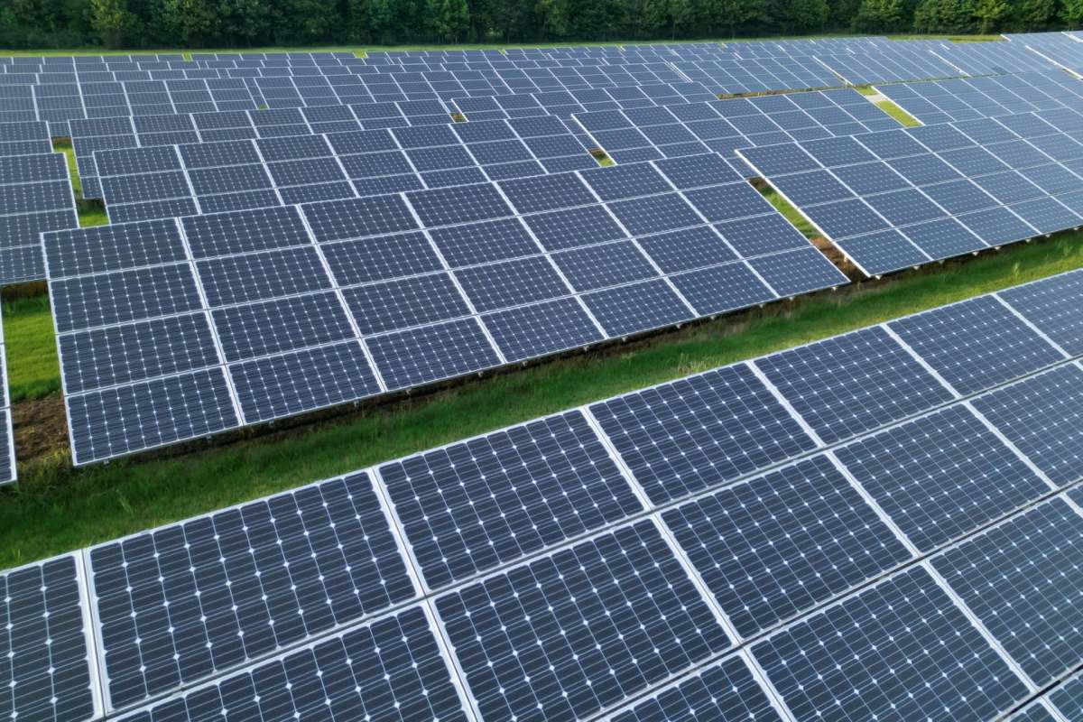 Aerial view of a large solar panel field with numerous panels arranged in rows on green grass.