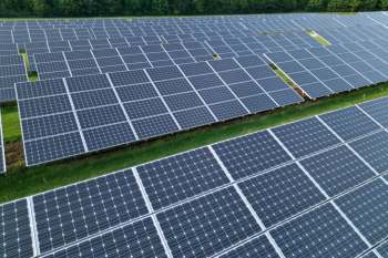 Aerial view of a large solar panel field with numerous panels arranged in rows on green grass.