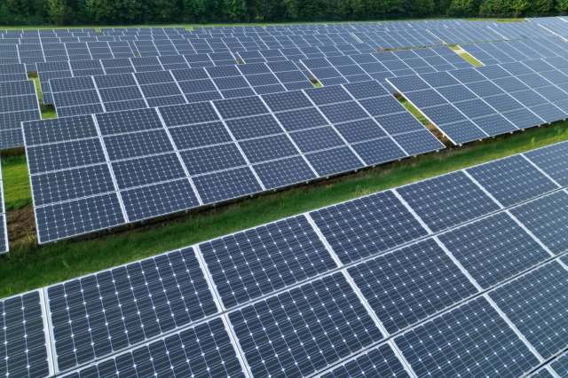 Aerial view of a large solar panel field with numerous panels arranged in rows on green grass.