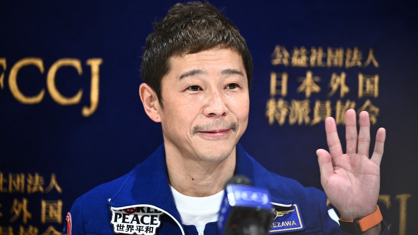 Japanese billionaire Yusaku Maezawa in a blue jumpsuit smiling and waving during a press conference.