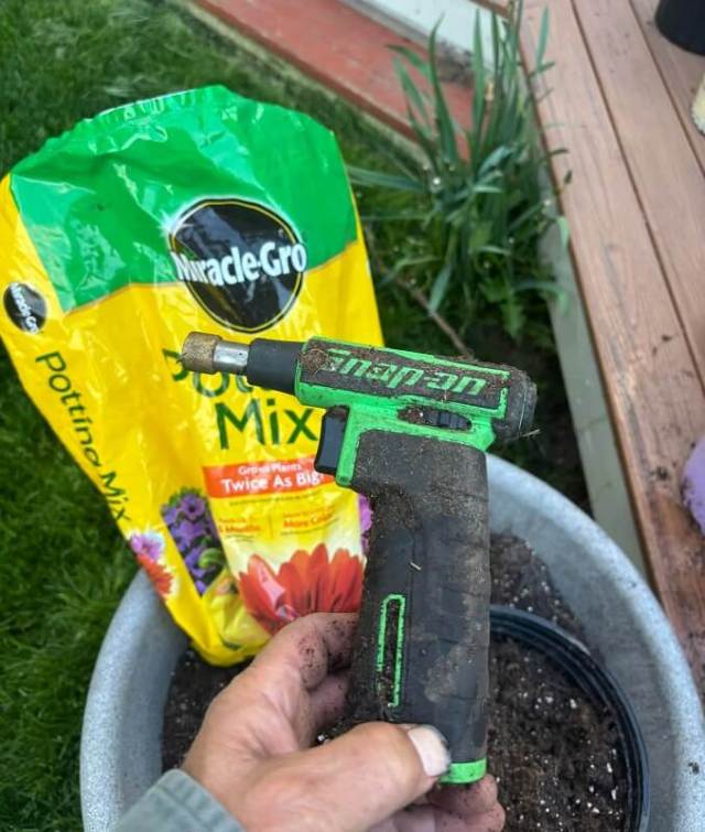 A close-up of a hand holding a dirty gardening tool with a yellow potting mix bag in the background.
