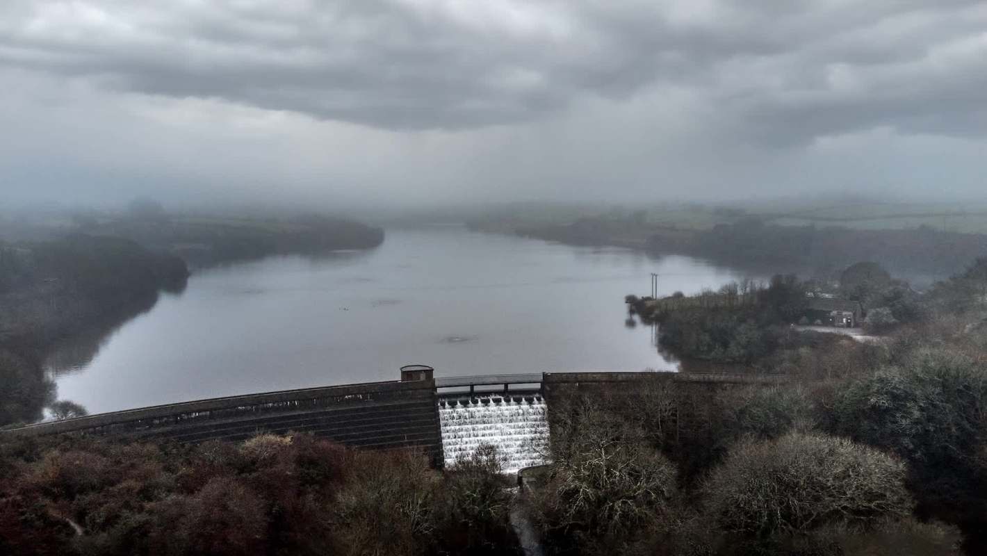 A misty landscape featuring a reservoir, a dam with flowing water, and a cloudy sky.