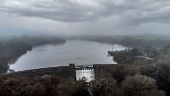 A misty landscape featuring a reservoir, a dam with flowing water, and a cloudy sky.