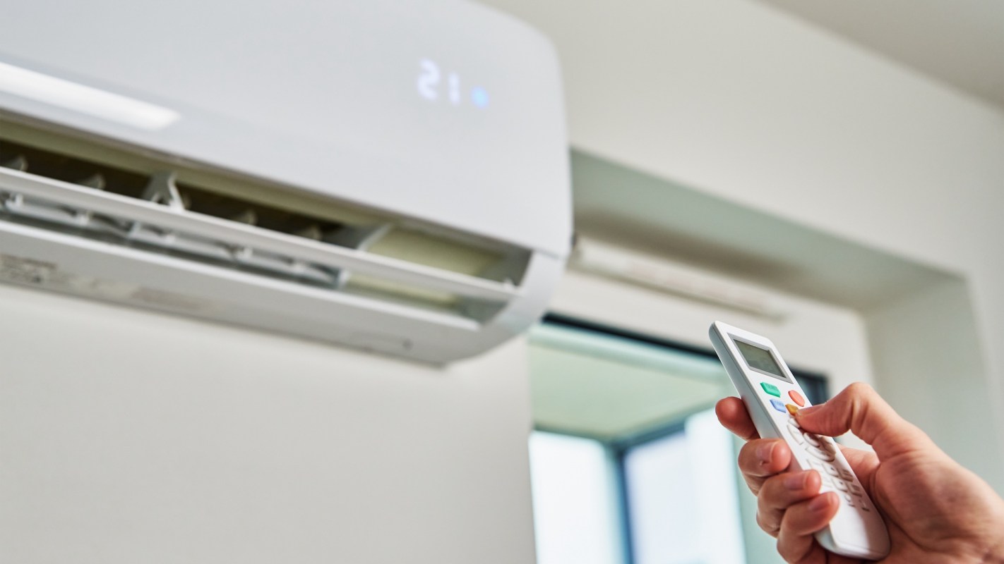 A hand holds a remote control in front of an air conditioning unit displaying a temperature of 21 degrees.