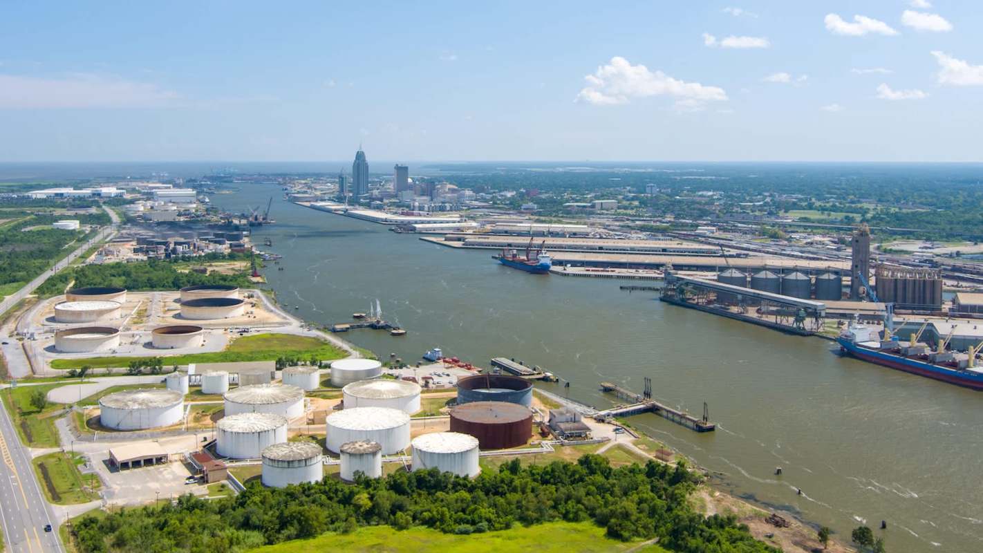 An aerial view of a busy industrial port with storage tanks, ships, and city skyline under a clear sky.