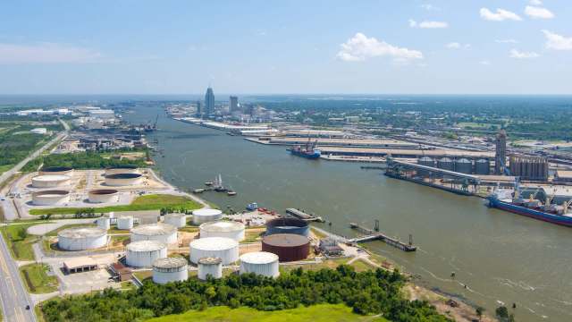 An aerial view of a busy industrial port with storage tanks, ships, and city skyline under a clear sky.
