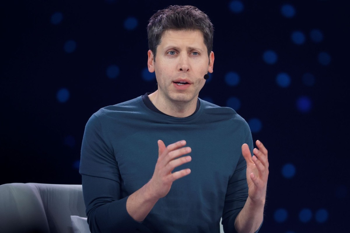Sam Altman gestures while speaking into a microphone on a stage with a blurred dark background.