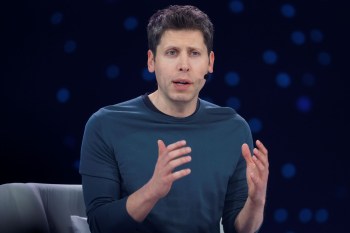 Sam Altman gestures while speaking into a microphone on a stage with a blurred dark background.
