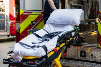 A stretcher with white bedding is positioned near the open doors of an ambulance.