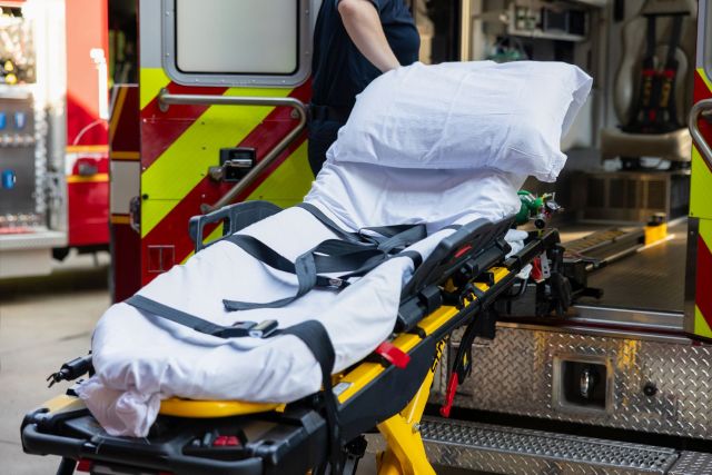 A stretcher with white bedding is positioned near the open doors of an ambulance.
