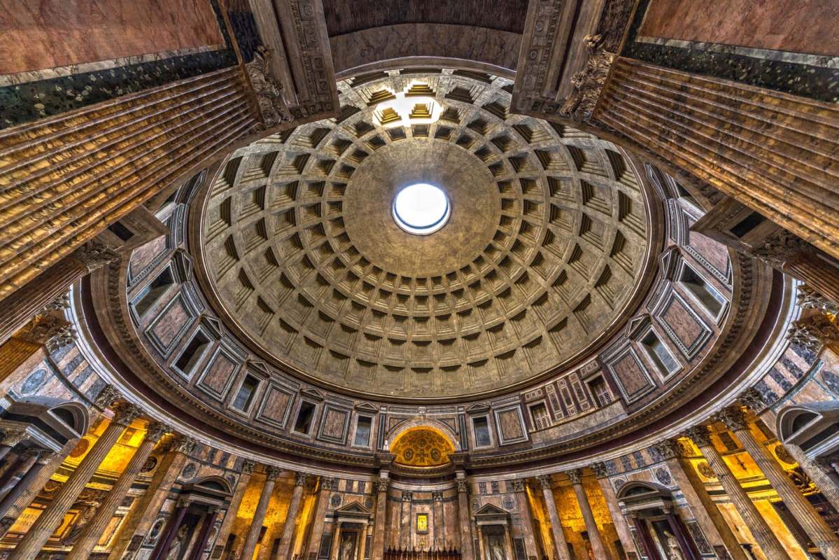 The intricate dome of a grand ancient building features a central oculus and ornate columns surrounding the interior.