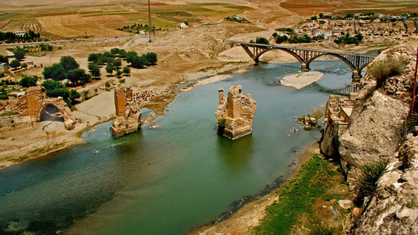 An aerial view of ruins along a riverbank with a bridge connecting both sides.