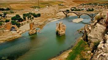 An aerial view of ruins along a riverbank with a bridge connecting both sides.