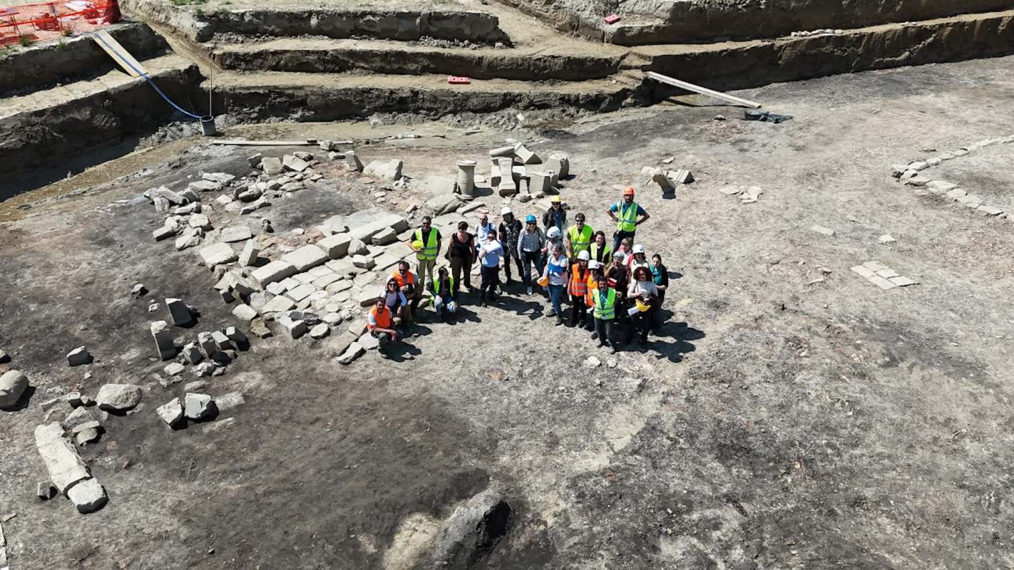 A group of archaeologists poses together at a dig site surrounded by stone structures and excavation areas.