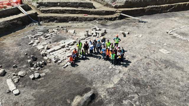 A group of archaeologists poses together at a dig site surrounded by stone structures and excavation areas.
