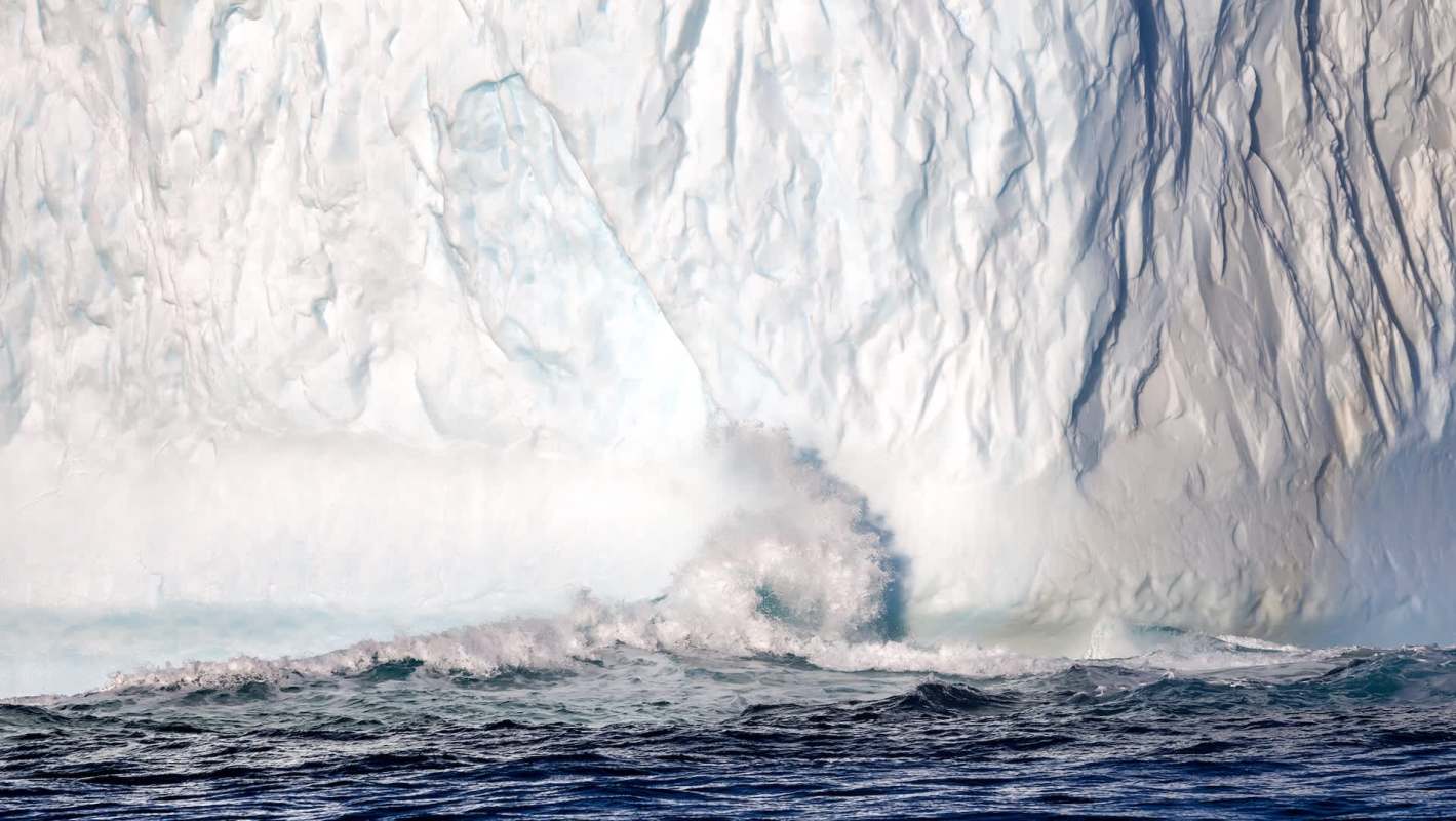 A large iceberg glistens in sunlight as waves crash against its base in deep blue water.