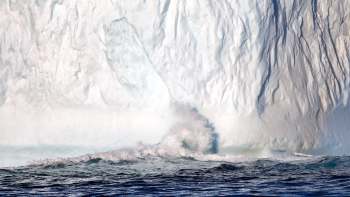 A large iceberg glistens in sunlight as waves crash against its base in deep blue water.