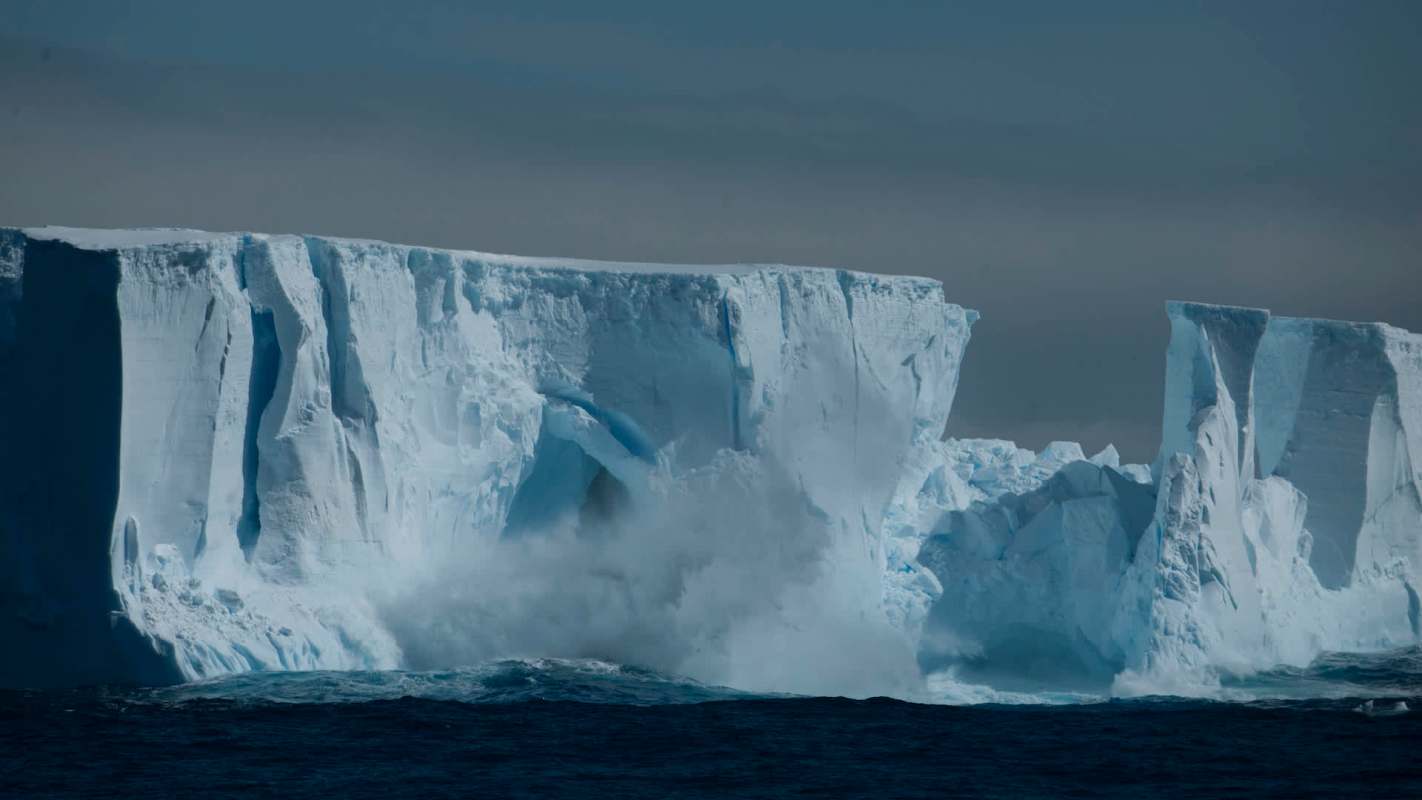 A massive iceberg breaks apart along the shoreline in the antarctic, sending waves crashing into the ocean.