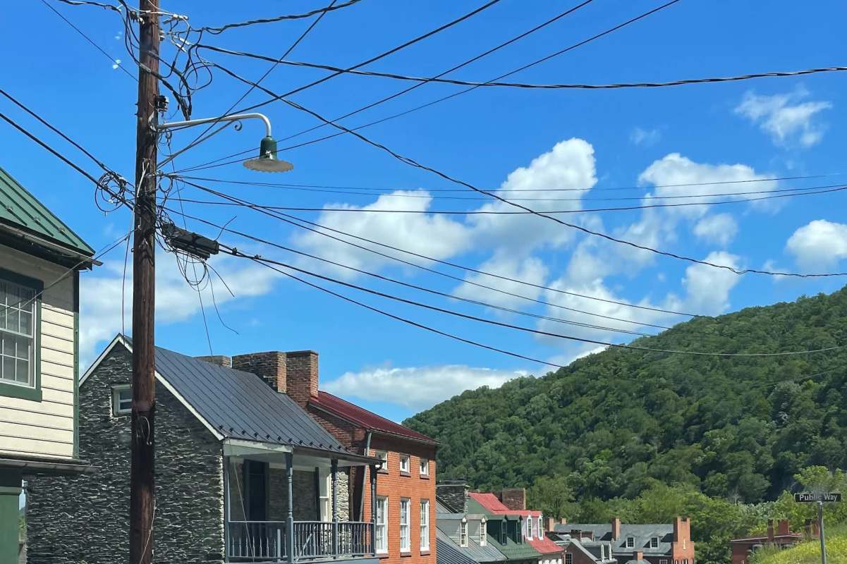 A street view showcasing vintage homes, power lines, and a green hillside under a bright blue sky with clouds.