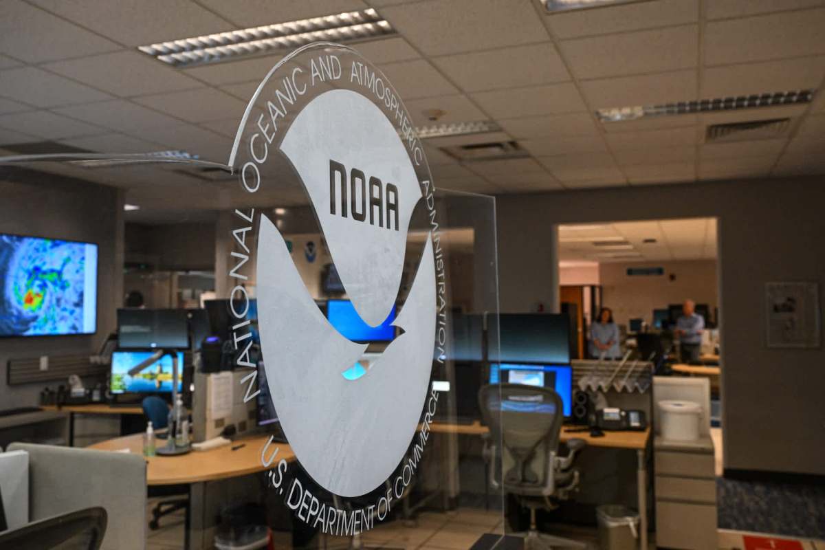 The NOAA logo is displayed on glass, with various computer monitors and staff visible in the background of an office setting.