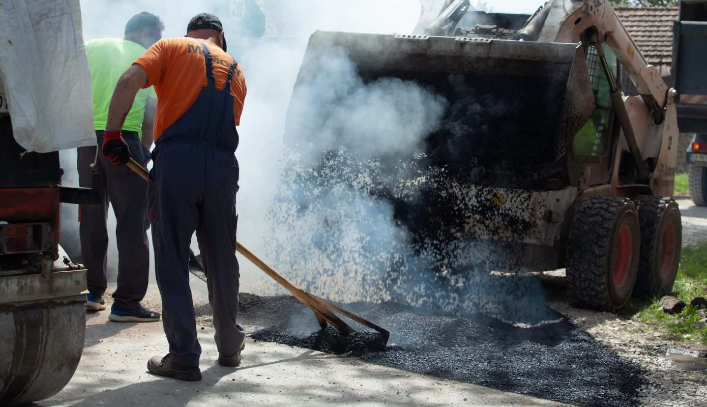 Workers are laying asphalt while smoke rises from a dump truck on a construction site.