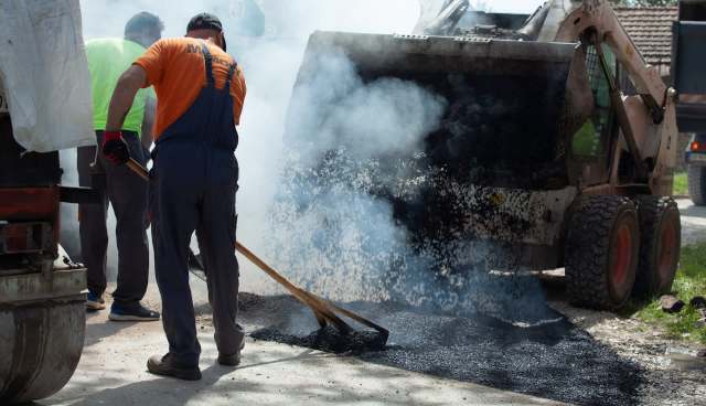 Workers are laying asphalt while smoke rises from a dump truck on a construction site.