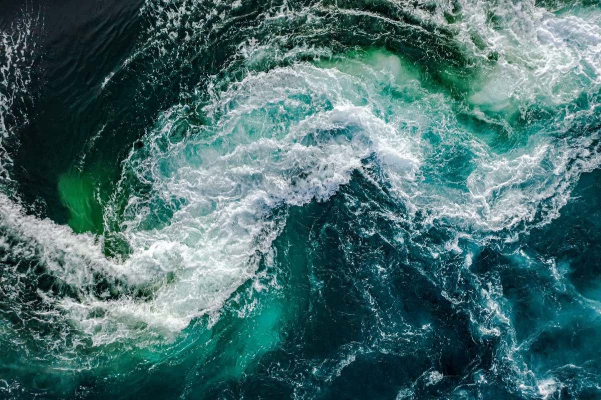 Aerial view of turbulent ocean water with swirling waves and varying shades of blue and green.