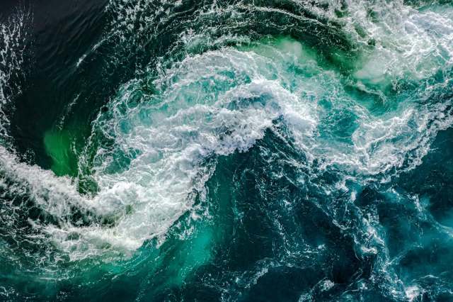Aerial view of turbulent ocean water with swirling waves and varying shades of blue and green.