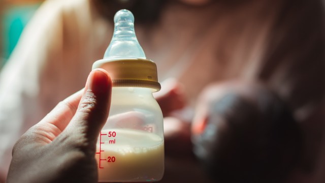 A hand holds a baby bottle filled with milk, with a soft-focused background of a caregiver and an infant.