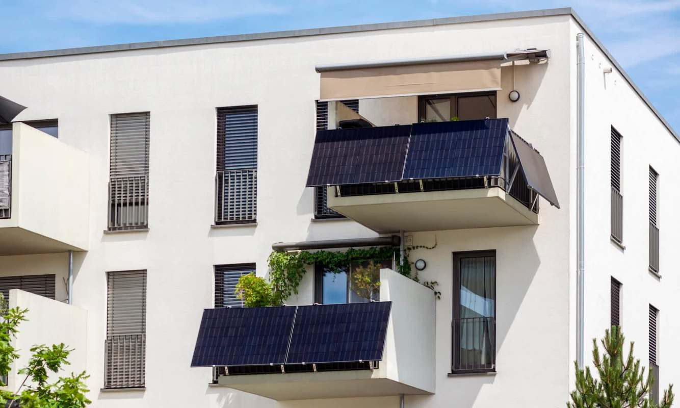 A modern apartment building with balconies featuring solar panels and greenery under a clear blue sky.