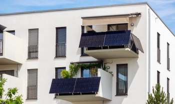 A modern apartment building with balconies featuring solar panels and greenery under a clear blue sky.