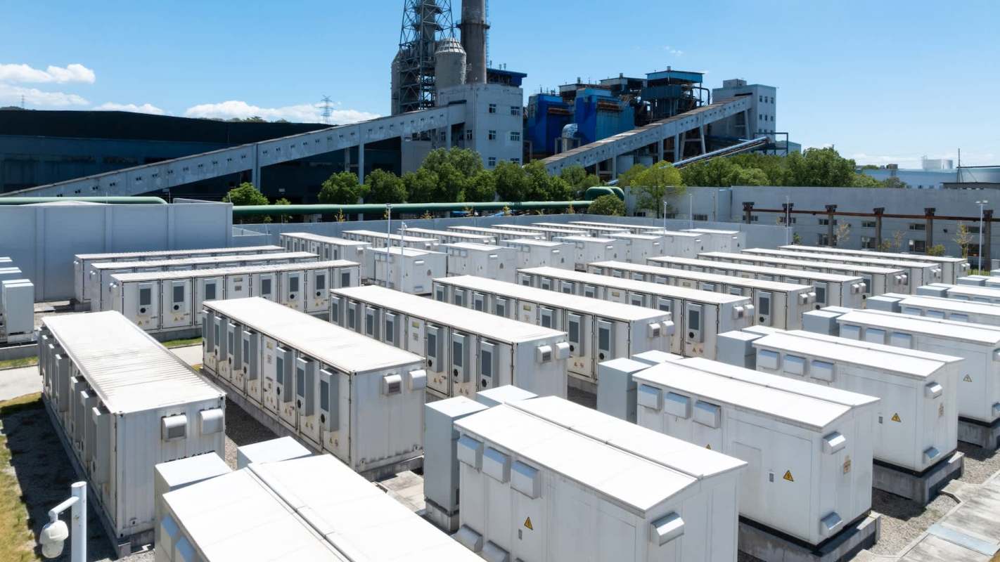 Aerial view of a battery storage installation with numerous white energy containers and a power plant in the background.
