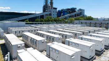 Aerial view of a battery storage installation with numerous white energy containers and a power plant in the background.