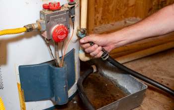 A hand holds a hose connected to a gas water heater, with various valves and pipes visible in the background.