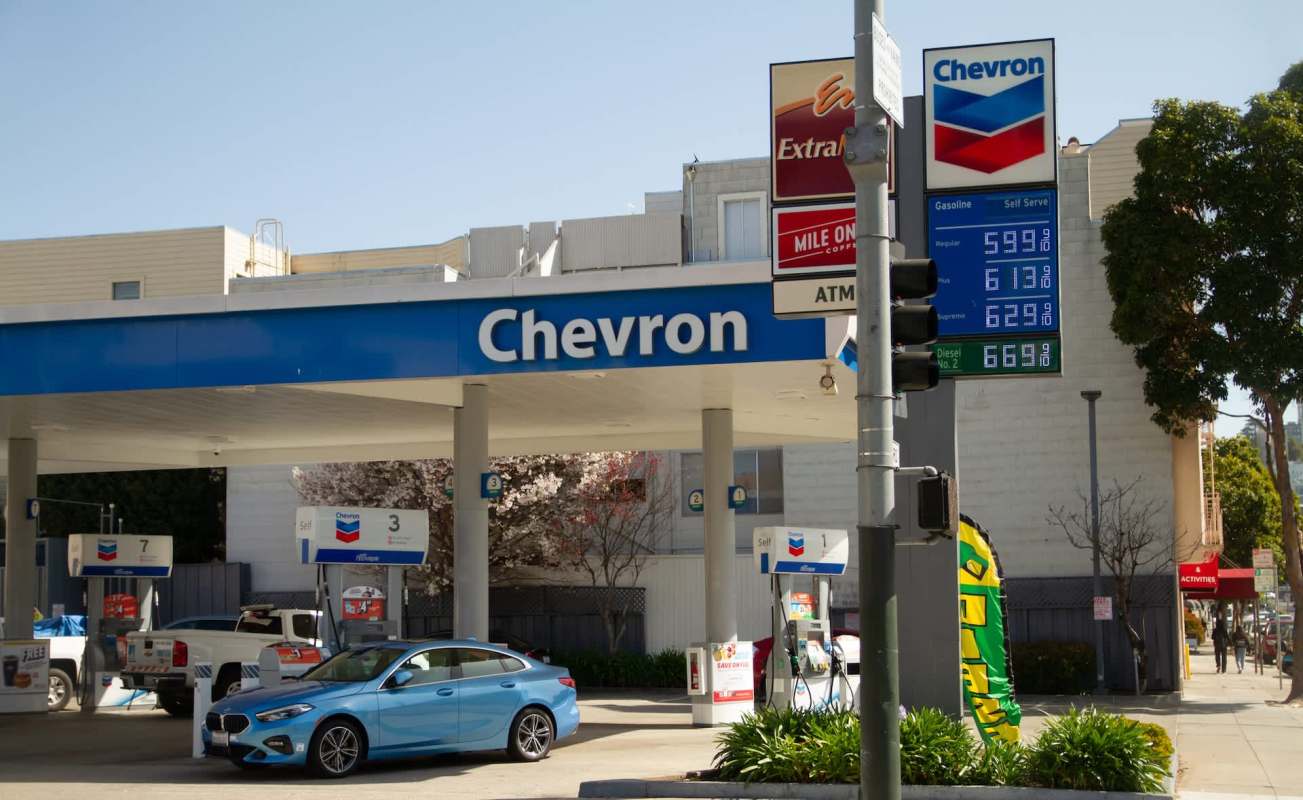 A Chevron gas station with prices displayed and a light blue car parked at the pump.