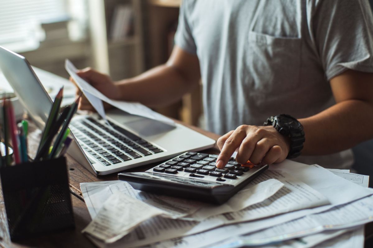 A person using a calculator while working on a laptop surrounded by papers on a wooden desk.
