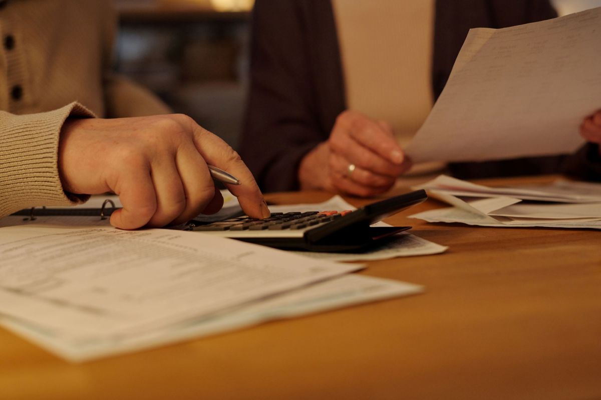 Two hands work on a calculator amidst scattered papers on a wooden table, suggesting a financial task.