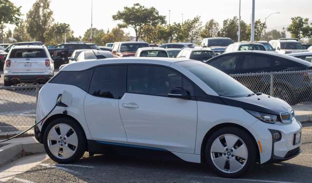 A white electric car is parked and charging in a parking lot surrounded by other vehicles.