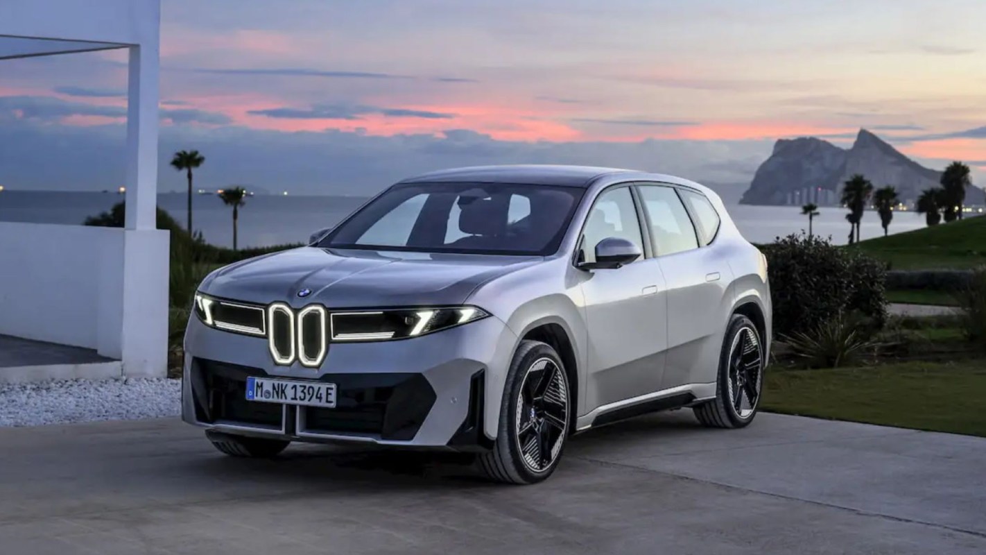 A silver BMW SUV parked on a driveway with a coastal view and mountains in the background at sunset.