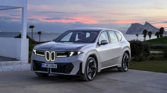 A silver BMW SUV parked on a driveway with a coastal view and mountains in the background at sunset.