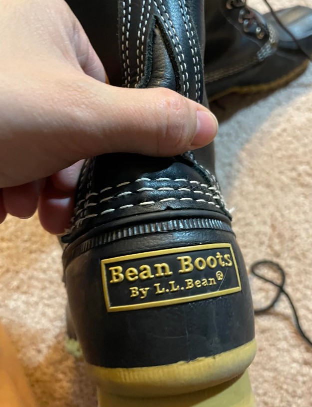 A hand shows a black leather boot, labeled Bean Boots By L.L.Bean, to the camera.