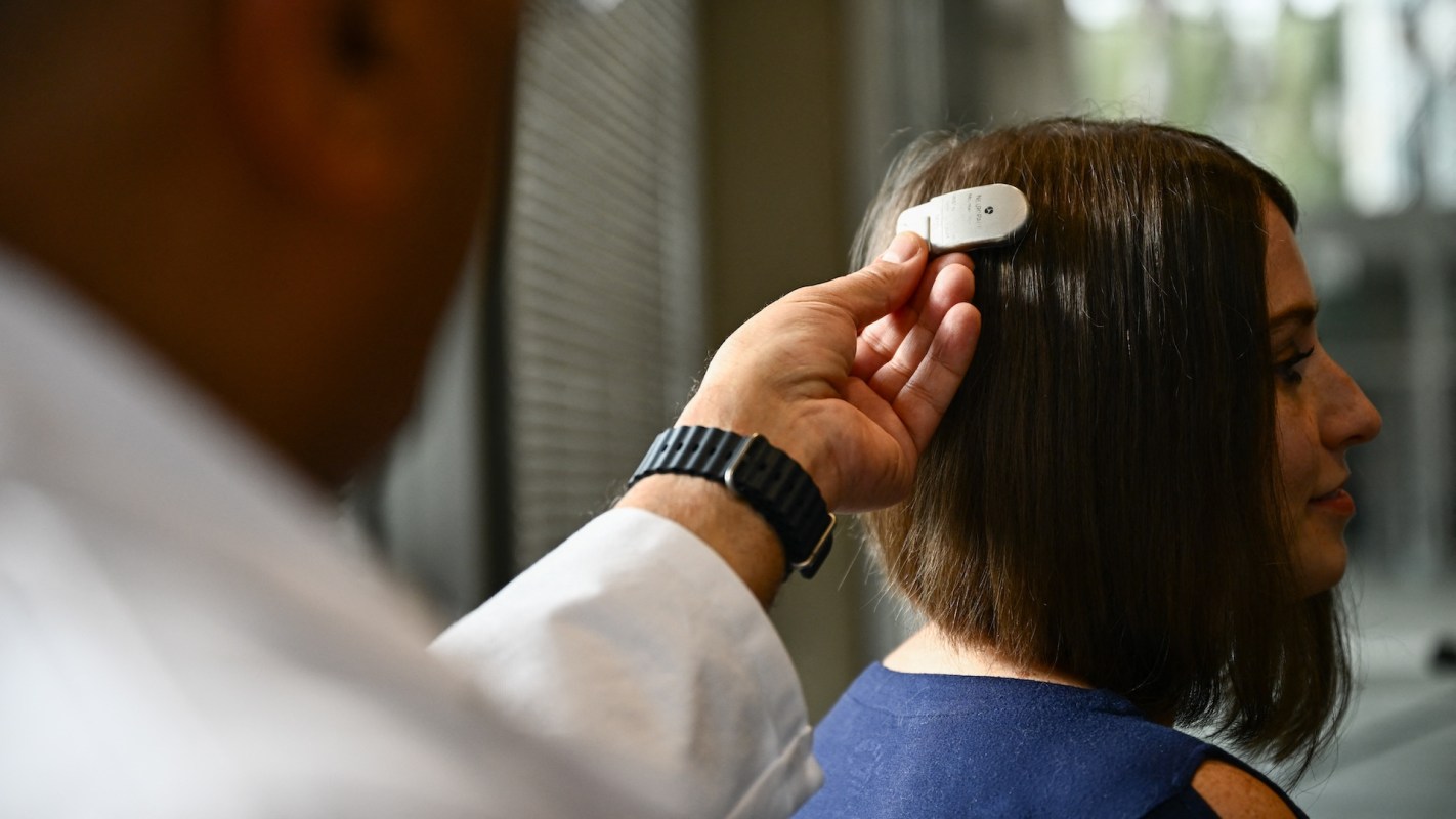 A person in a lab coat is using a device on the hair of a woman with short brown hair.