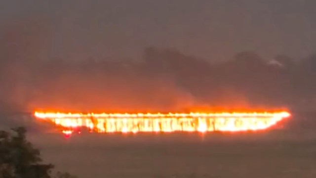 A long, fiery line on the horizon, surrounded by dark smoke from a bridge burning.