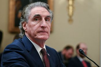 Doug Burgum sitting at a desk with a microphone and blurred figures in the background.