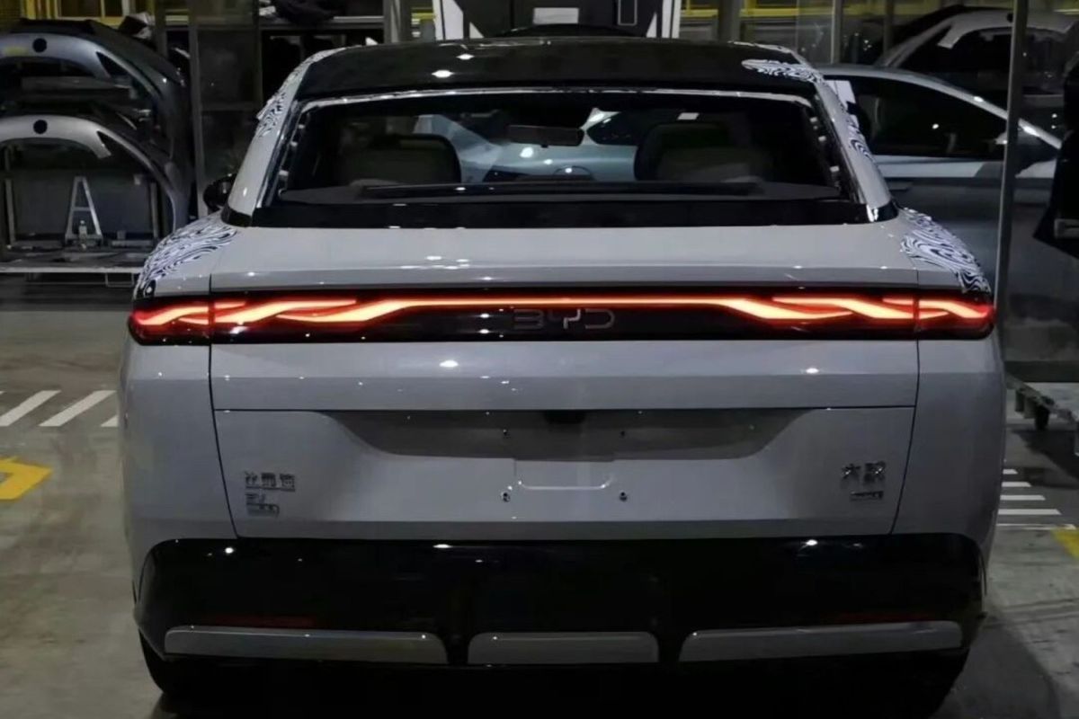 A rear view of a white BYD electric car with sleek taillights and partial camouflage, surrounded by an industrial setting.