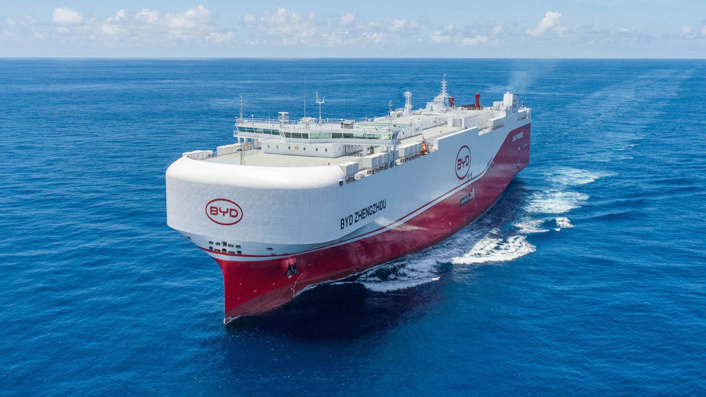 A large cargo ship with a white and red hull sails through deep blue ocean waters under a clear sky.