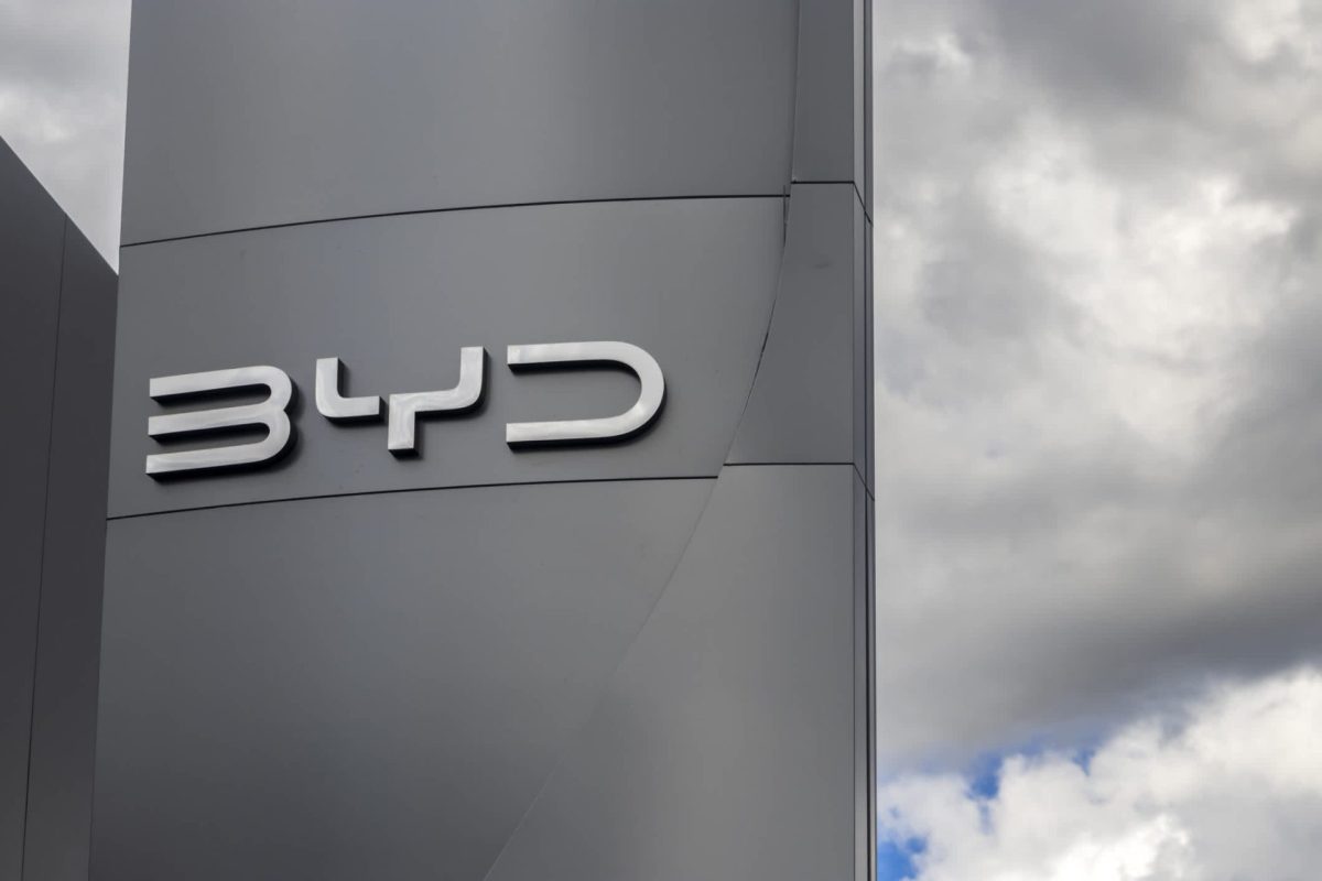 A close-up of a sleek silver BYD logo against a gray background and cloudy sky.